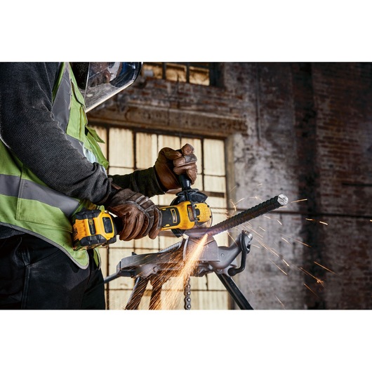 A person wearing protective gloves and a safety visor is using a yellow DEWALT cordless angle grinder to cut through a metal rod, with sparks flying from the cutting area. The work is being done indoors, in front of a brick wall with large windows.