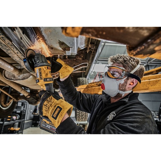 A person wearing safety equipment uses a DEWALT 18V XR FLEXVOLT cordless angle grinder to cut metal underneath a vehicle in a workshop. Sparks are visible where the tool is in contact with the metal.