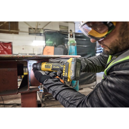 A person wearing gloves uses a DEWALT DCG420N cordless power tool to grind or polish metal inside an industrial workshop.