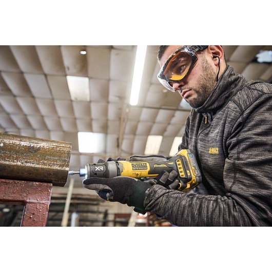 A person wearing a DEWALT jacket and gloves is using a DEWALT cordless rotary tool (model DCG420N) to work on a piece of metal in an industrial workshop.