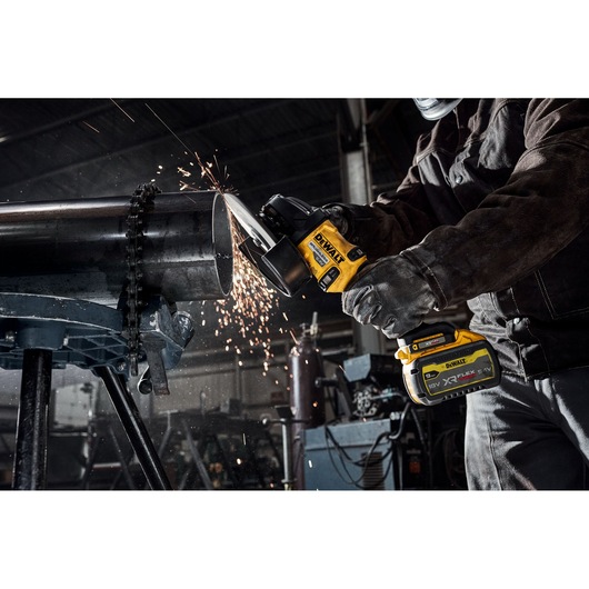 A worker uses a DEWALT cordless angle grinder to cut a metal pipe, with sparks flying in an industrial workshop setting.