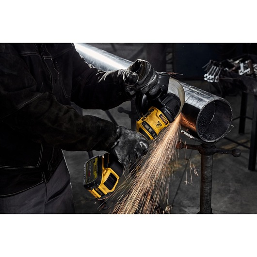 A person wearing protective gloves uses a yellow DEWALT DCG440N cordless grinder to cut a metal pipe, with bright sparks flying from the grinding wheel.
