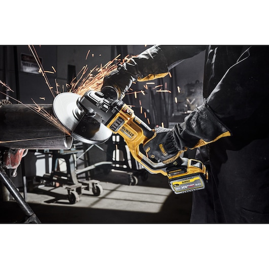 A person wearing protective gloves and dark clothing is using a yellow DEWALT cordless grinder to cut through a metal pipe, producing sparks, in an industrial workshop setting.