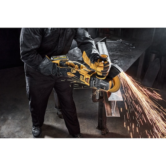 A person wearing protective gloves and clothing is using a DEWALT battery-powered angle grinder to cut a metal piece, producing bright sparks in a workshop setting.