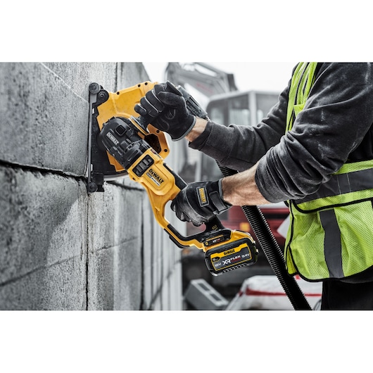 A person wearing work gloves and a safety vest using a DEWALT cordless wall chaser tool to cut grooves into a concrete block wall at a construction site.