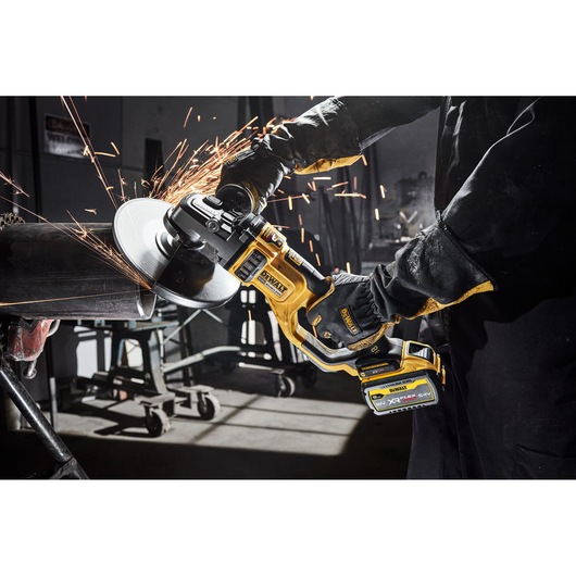 A person wearing protective gloves and clothing is using a DEWALT cordless angle grinder to cut a metal pipe, producing sparks in an industrial workshop setting.