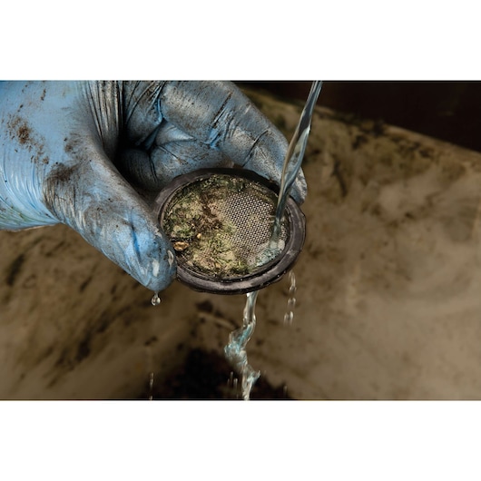A person wearing a dirty blue glove is cleaning a gritty, circular metal filter under running water.