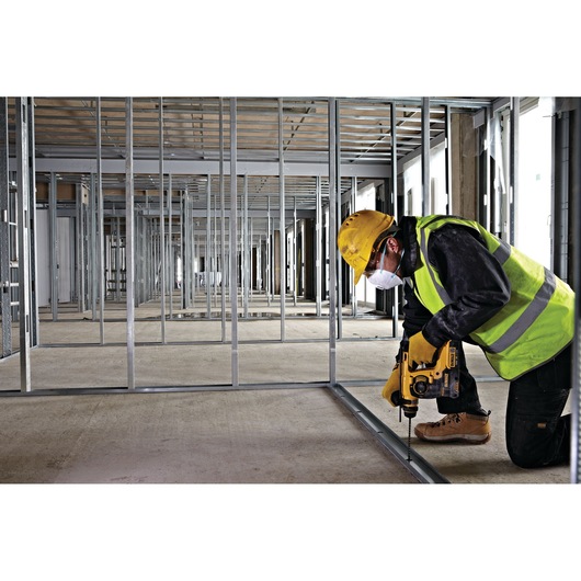 Worker with yellow drill installs metal frame inside a building under construction, wearing safety gear.