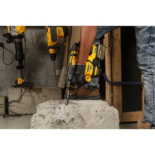 A person using a DEWALT DCH333NT cordless rotary hammer to drill into a concrete block. The worker is wearing gloves and work boots, and other DEWALT power tools are visible in the background.