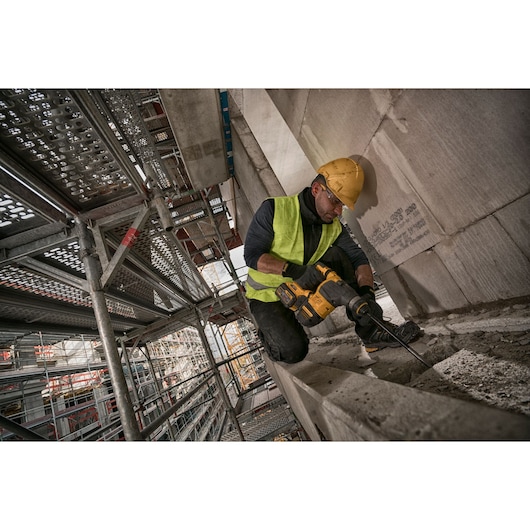 Worker using a DEWALT rotary hammer drill on a construction site with scaffolding in the background.