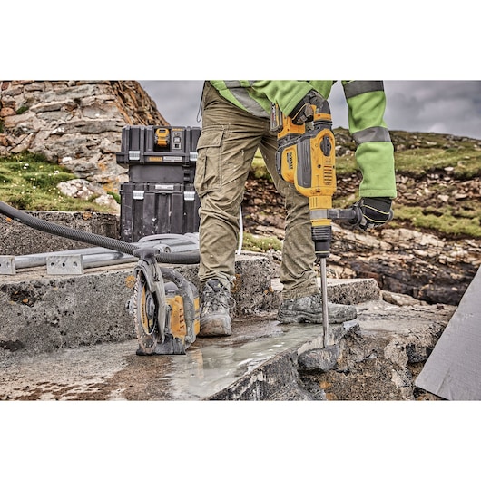 Person using a yellow DEWALT rotary hammer drill to break concrete outdoors, with additional DEWALT tools and toolboxes visible in the background.