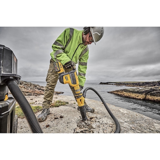 A person wearing a green safety jacket, gloves, and a helmet is using a yellow DEWALT rotary hammer drill to break concrete outdoors near a body of water. A vacuum hose is attached to the tool to collect dust.
