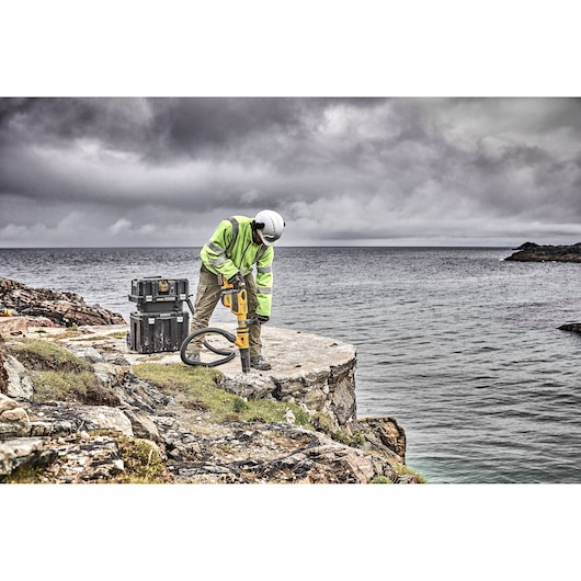 A worker in a high-visibility jacket and helmet is using a yellow DEWALT demolition tool on a rocky cliff above the ocean, with DEWALT storage boxes nearby. The sky is cloudy.