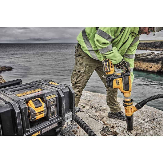 A person in a high-visibility jacket operates a DEWALT power tool to drill into a stone surface outdoors near water, with a DEWALT tool case and battery pack in the foreground.