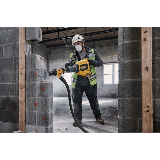 A construction worker wearing safety gear uses a DEWALT rotary hammer to drill into a concrete block wall inside a building under construction. The worker is holding the tool, which is connected to a dust extraction hose.
