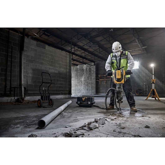 A construction worker wearing a safety helmet and reflective vest uses a DEWALT demolition tool to break concrete in an industrial setting. Nearby are a pipe, a tool case, a dolly, and a standing work light.