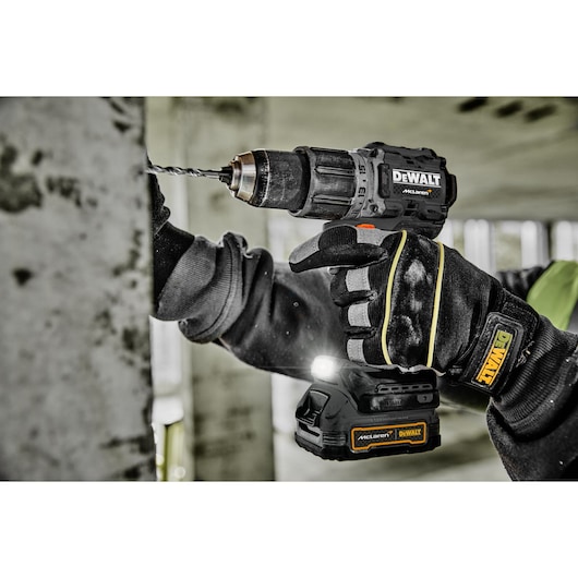 A person wearing protective gloves uses a DEWALT cordless drill to drill into a concrete wall. The drill has a McLaren branded battery pack and an LED light. The scene appears to be in a construction site.