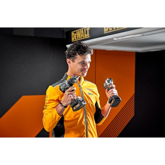 Person wearing a yellow DEWALT and McLaren-branded jacket holding two cordless DEWALT power tools, standing indoors with a DEWALT logo in the background. The person's face is blurred.