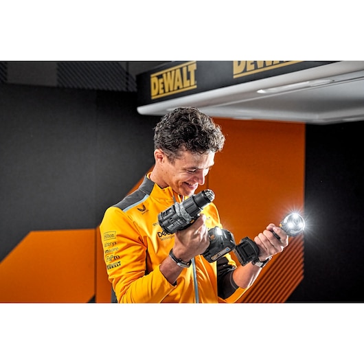 A person in a yellow and black branded jacket is holding two DEWALT power tools, a drill and a flashlight, inside a room with DEWALT signs. The person's face is blurred.