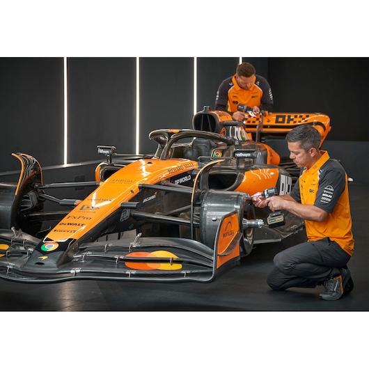 Two technicians wearing orange shirts are working on an orange Formula 1 race car inside a brightly lit garage. One technician is using a DEWALT power tool on the car's front wing, while the other is leaning over the rear section of the vehicle.