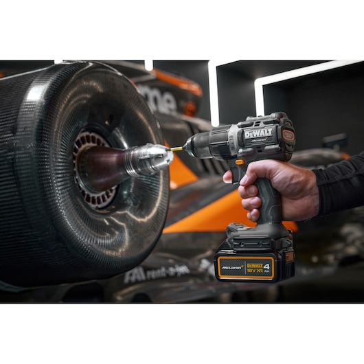 A person using a DEWALT cordless drill on the wheel of a racing car. The drill is labeled with McLaren and DEWALT branding, and the car in the background features orange and black colors.