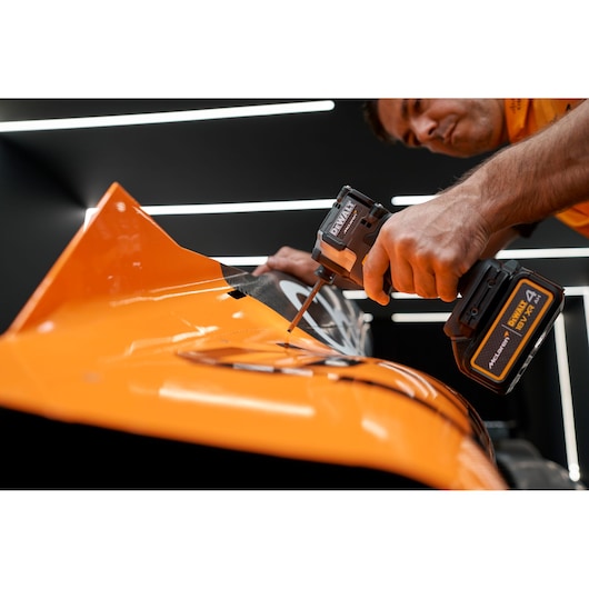A person uses a DEWALT cordless power tool with a 4Ah XR lithium-ion battery to fasten a screw on an orange metal surface, likely part of machinery or equipment.