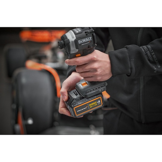 Close-up of a person holding a DEWALT 18V XR cordless power tool with a McLaren-branded 4Ah battery pack. The background shows a blurred racing car with orange accents.