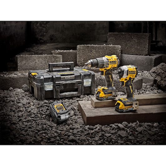 A set of DEWALT power tools including a cordless drill, an impact driver, a charger, and a toolbox, displayed on a construction site with concrete blocks and rubble.