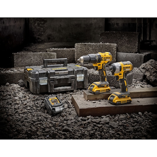 Two DEWALT cordless power tools, a drill and an impact driver, are displayed on a construction site among rubble and concrete blocks. Next to them are a DEWALT toolbox and a battery charger.