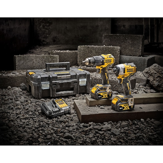 A DEWALT tool kit set is displayed on a construction site, featuring two yellow and black cordless power tools, a storage case, and a battery charger. The tools are set on wooden blocks amid gravel and concrete bricks.