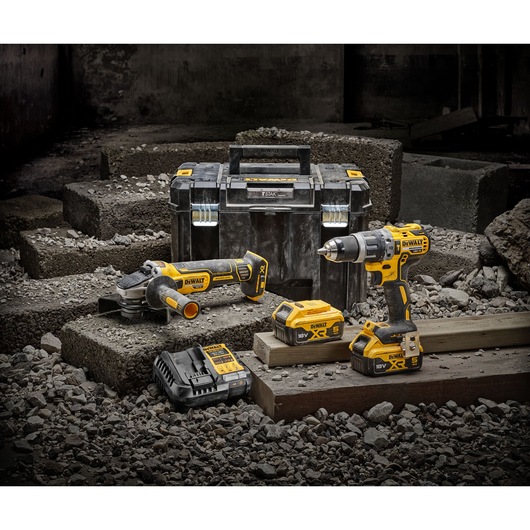 A DEWALT tool set featuring a cordless drill, an angle grinder, two 18V batteries, a battery charger, and a durable black and yellow carrying case, displayed on a construction site with concrete blocks and rubble.