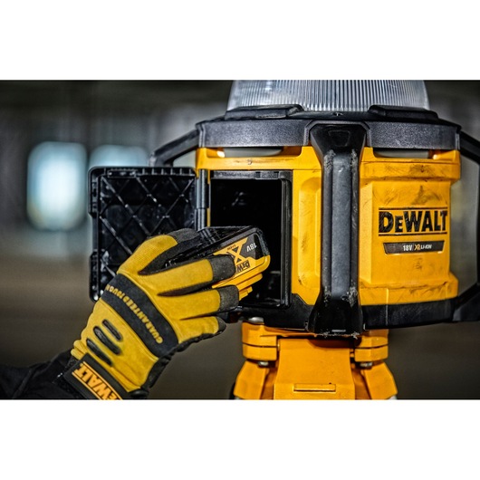 A close-up of a gloved hand inserting a DEWALT 18V battery into a yellow DEWALT portable work light, model DCL074, with the battery compartment open.
