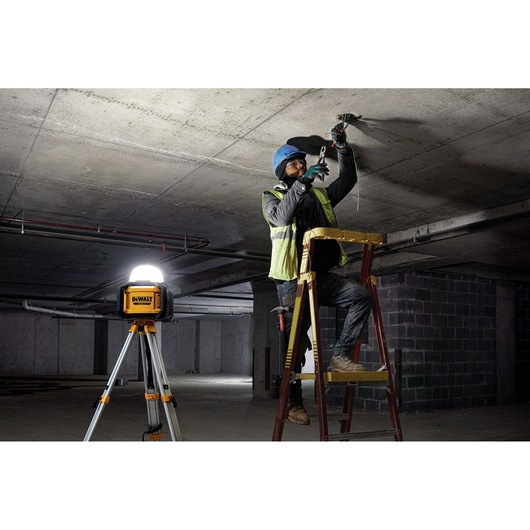 A construction worker wearing a safety vest and helmet stands on a ladder, working on wiring attached to a concrete ceiling. A DEWALT DCL074 site light is positioned on a tripod nearby, illuminating the workspace in an unfinished building.