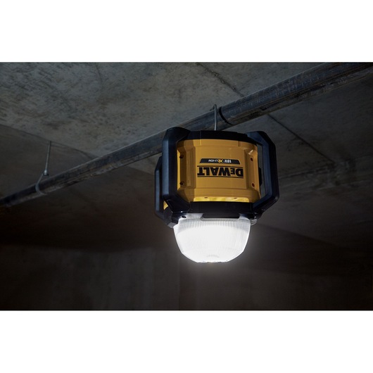 A DEWALT DCL074 LED work light is hanging from a metal pipe on a concrete ceiling, providing bright illumination in an industrial or construction setting.