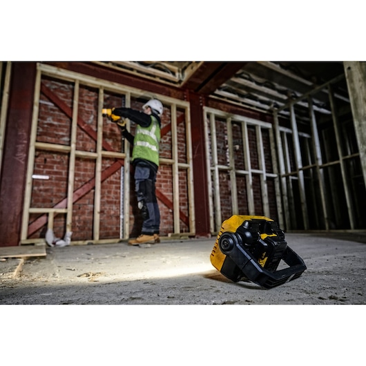 A DEWALT portable work light (SKU: DCL077) sits on the floor of a construction site, illuminating the area. In the background, a construction worker wearing safety gear is working near a wooden frame against a brick wall.