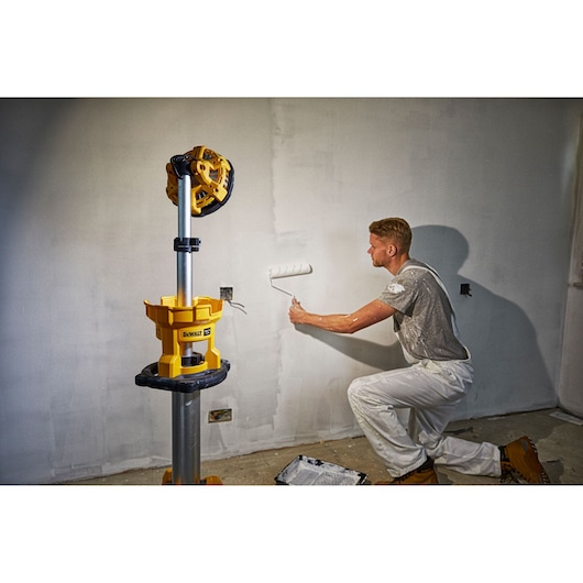 A person painting a wall with a roller while a DEWALT DCL079 standing LED work light is illuminated beside them. The scene suggests a renovation or construction environment.