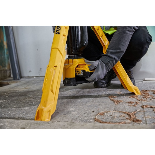 A close-up of a person wearing gloves, installing a battery into a yellow DEWALT DCL079 tripod work light set up on a concrete floor.