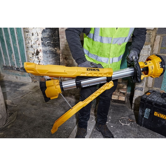 A person wearing a reflective vest handles a DEWALT DCL079 tripod-style portable work light in a construction or renovation setting. The yellow and black light is being unfolded and is positioned in front of a brick wall and toolbox.