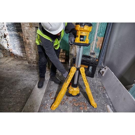 A worker wearing a safety helmet and vest adjusts a yellow DEWALT DCL079 tripod work light in an industrial or construction setting. There are debris and tools around the work area.