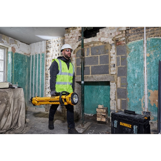 A person in safety gear carrying a yellow DEWALT portable LED area light (model DCL079) inside a construction site. There is a DEWALT toolbox on the floor and exposed brick and plaster walls in the background.