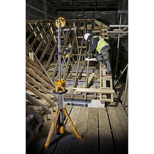 A DEWALT DCL079 tripod-mounted work light is positioned in the foreground, illuminating a construction site with wooden beams and scaffolding. A worker wearing a safety vest and helmet is operating tools on a platform.