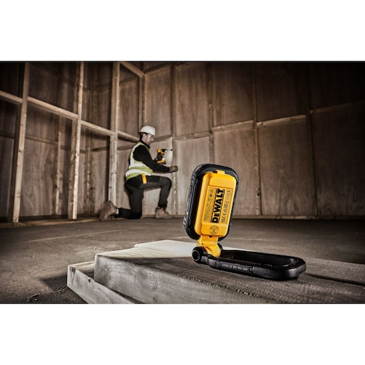 A DEWALT DCL182 portable work light is placed on a concrete surface in an unfinished room with wooden framing. In the background, a person wearing a safety vest is kneeling and using a cordless tool on the wall.