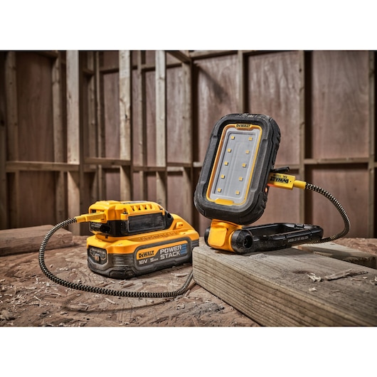 A DEWALT DCL182 portable LED work light connected to a DEWALT POWERSTACK 18V 5Ah battery, placed on a wooden surface in a construction site setting.