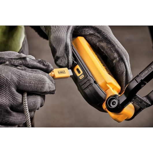Close-up of gloved hands plugging a DEWALT-branded USB cable into the charging port of a yellow DEWALT device, likely a portable work light.