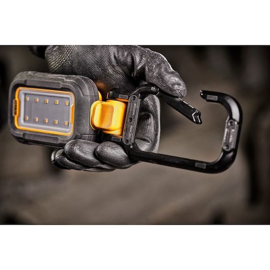 A close-up of a gloved hand holding a DEWALT portable LED work light with a built-in carabiner clip for easy attachment.