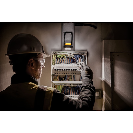A person wearing a safety helmet and gloves is working on an electrical panel, illuminated by a DEWALT work light mounted above the panel.