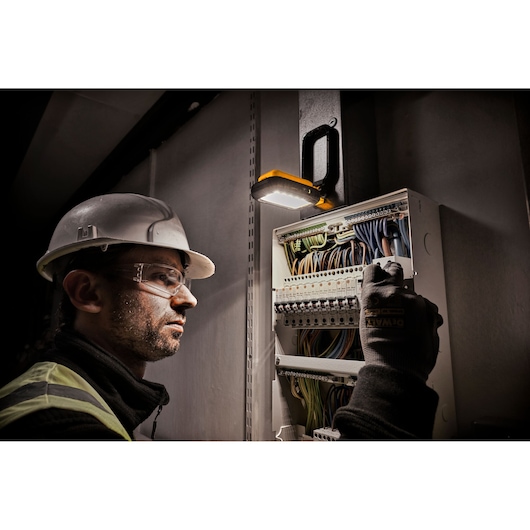 A person wearing a safety helmet and gloves is working on an electrical panel, illuminated by a DEWALT portable work light (SKU: DCL182) mounted above the panel.