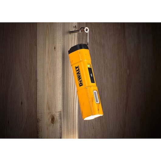 A yellow DEWALT DCL183 inspection light with a black top, hanging from a screw on a wooden wall, illuminating downward.