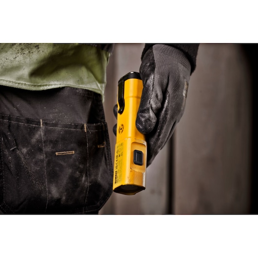 A close-up of a person wearing a work glove holding a yellow DEWALT DCL183 portable work light, standing next to a tool pouch.
