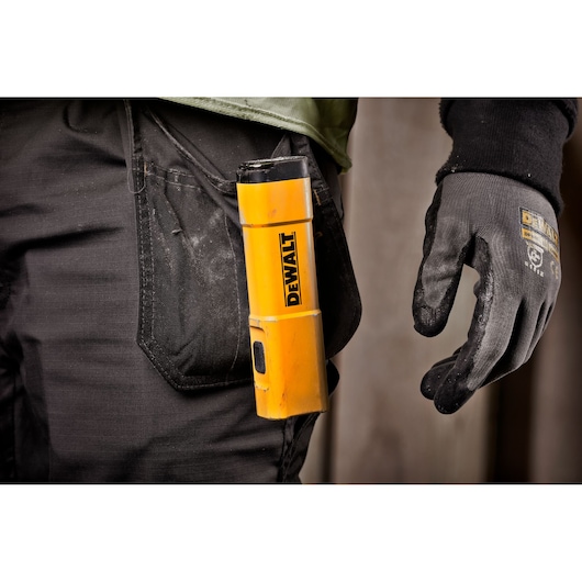 A close-up of a person wearing black work pants and gloves, with a yellow DEWALT DCL183 flashlight clipped to their pocket.